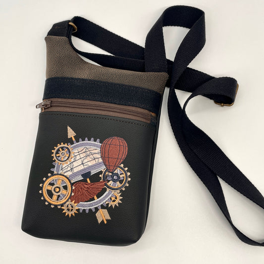 Pochette en simili noir brodé d'engrenages style steampunk. et de simili daim marron.  Elle a une poche zippée devant et une poche zippée au dos. Bandoulière en sangle noire réglable. Doublure imprimée avec des motifs engrenages assortis.